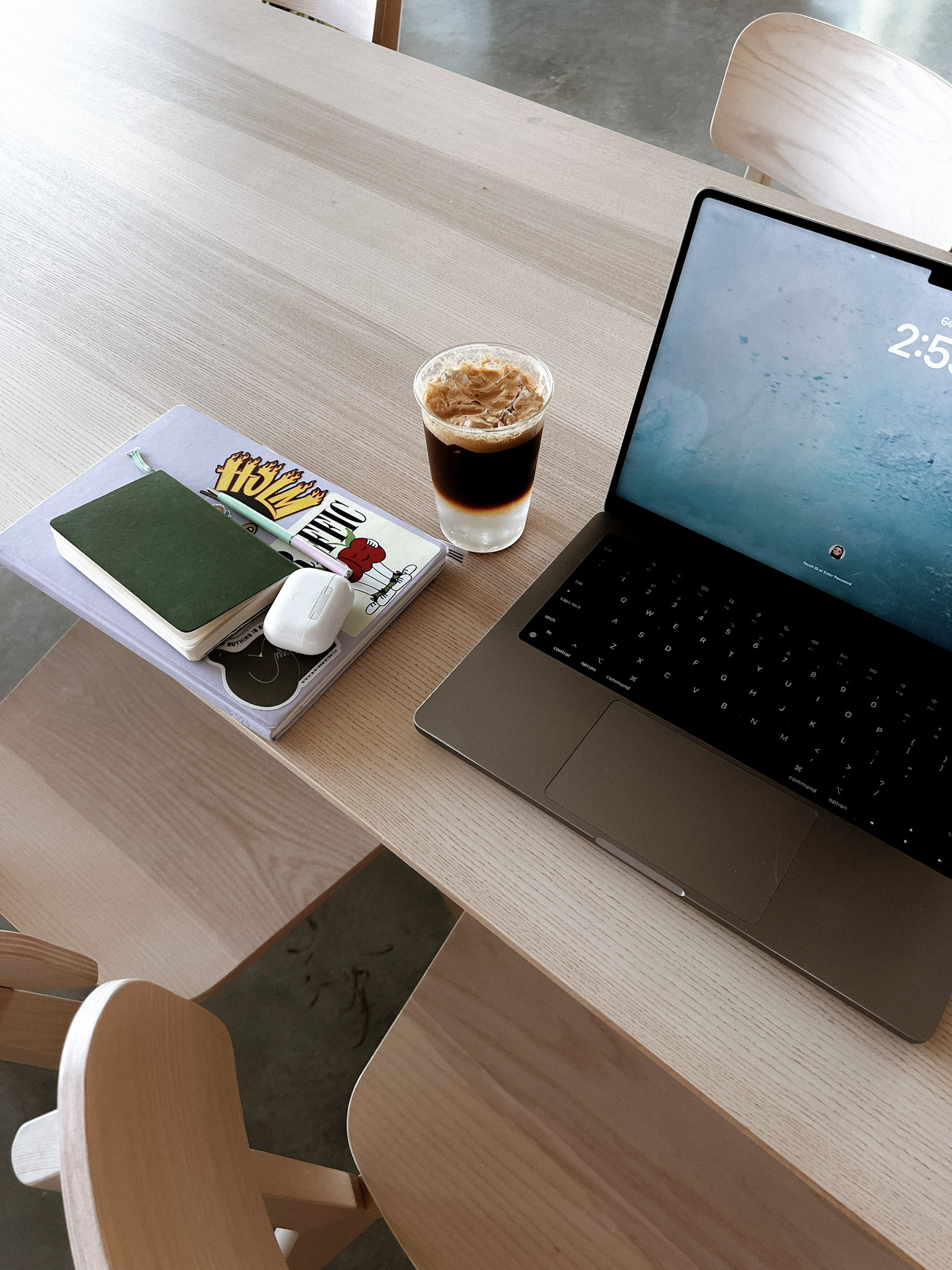 A minimalist workspace with a laptop, layered iced coffee, and stacked notebooks on a light wooden table, creating a clean and productive environment.